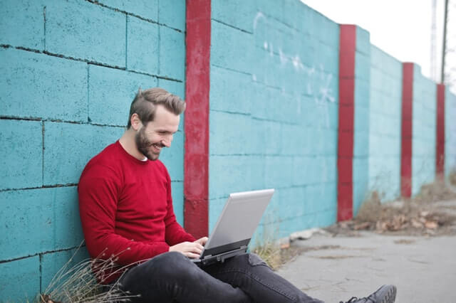 man leaning against wall with laptop and laughing 